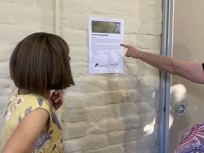 Person pointing to an A3 poster about an engagement method, with women watching to the side. 