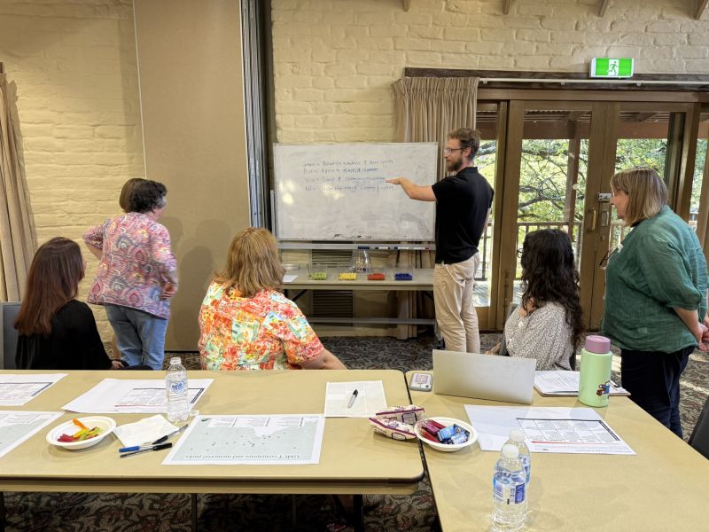 Standing man pointing at a whiteboard with four women standing and sitting near by watching.