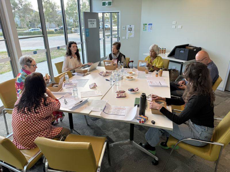 Seven people in deep discussion seated around a square table with A3 documents and snacks in front of them